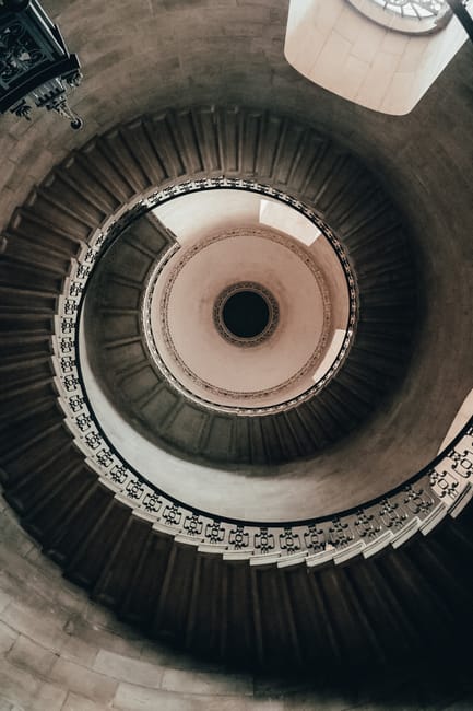 A monochrome photograph capturing a top-down view of a spiral staircase in a building, with concrete steps curving inward as they descend, bordered by a black metal railing featuring vertical baluster