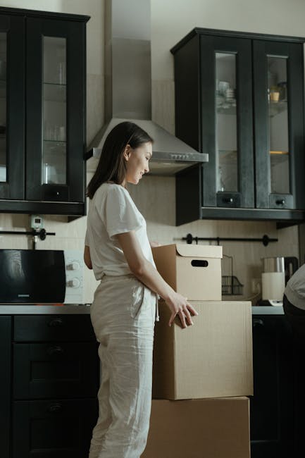 Interior scene during a house relocation showing two individuals engaged in packing and organizing moving boxes. A man with dark hair tied in a bun, wearing a dark grey t-shirt and beige trousers, is