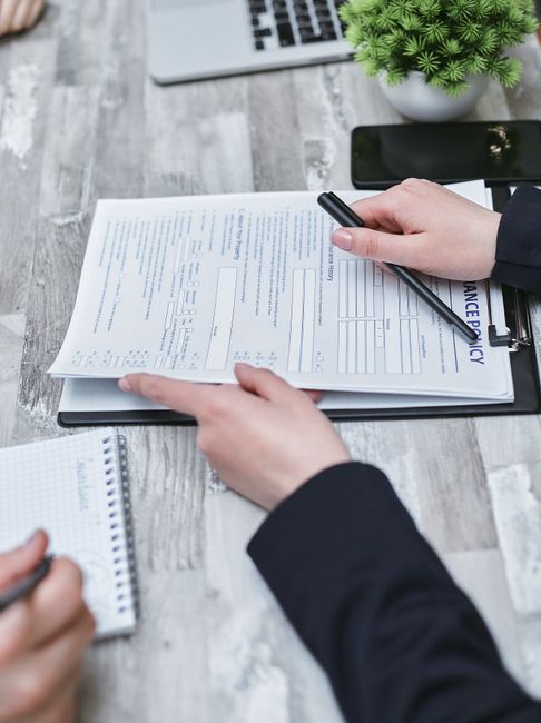 A person wearing a dark suit is seated at a grey wooden table, holding a pen and reviewing a document titled 'Insurance Policy' with detailed text and fields for information. On the table, there is a